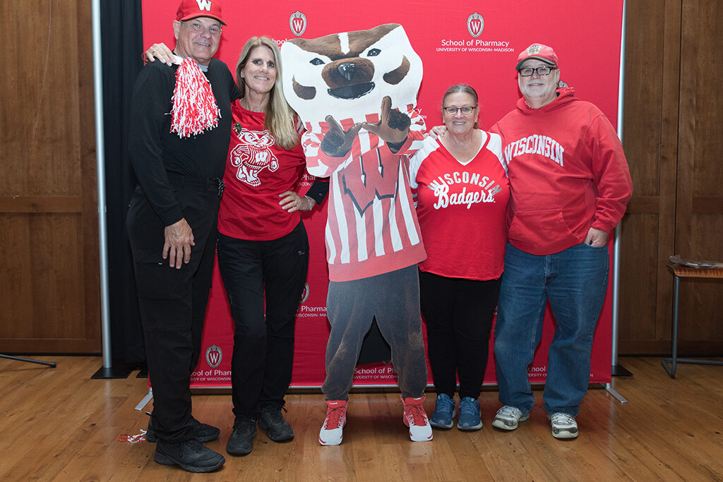 School of Pharmacy Alumni Tailgate Party 2023 Alumni pose with Bucky cardboard cutout in front of red School of Pharmacy logo background