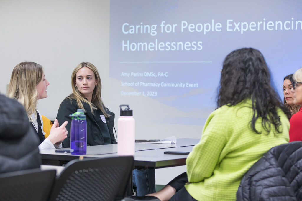 SoP Lecture: Cultivating Belonging in Pharmacy (December 1, 2023) PharmD students talking around a table, with a Caring for People Experiencing Homelessness presentation on the wall behind them.